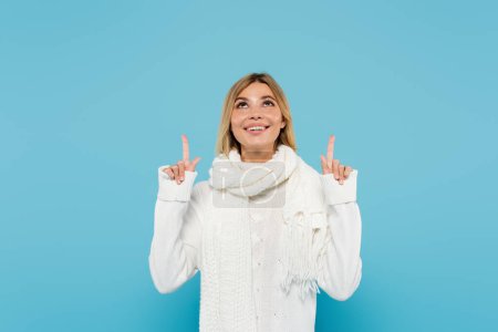 mujer rubia alegre en suéter blanco y bufanda apuntando hacia arriba con los dedos aislados en azul 
