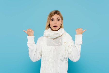 zdezorientowany blondynka kobieta w białym swetrze i szalik wskazując z kciuki odizolowane na niebieski 