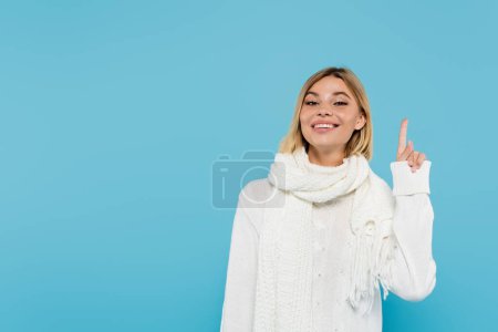 mujer rubia alegre en suéter blanco y bufanda apuntando hacia arriba con el dedo aislado en azul 