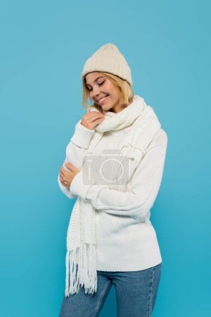 Photo for Happy blonde woman in white sweater and winter hat smiling isolated on blue - Royalty Free Image