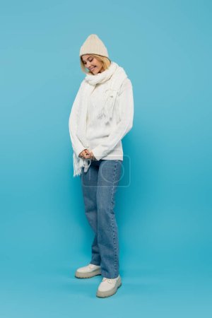Foto de Longitud completa de mujer rubia tímida en suéter blanco y sombrero de invierno sonriendo en azul - Imagen libre de derechos