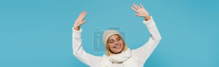 Photo for Positive blonde woman in white sweater and winter hat raising hands isolated on blue, banner - Royalty Free Image