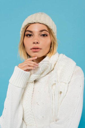 Téléchargez les photos : Portrait de jeune femme blonde en pull blanc, écharpe d'hiver et bonnet tricoté isolé sur bleu - en image libre de droit