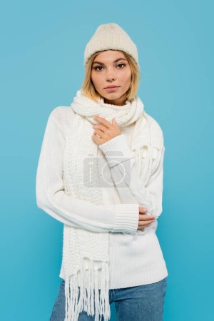 Photo for Portrait of young blonde woman in white sweater, scarf and knitted hat looking at camera isolated on blue - Royalty Free Image