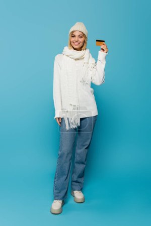 volle Länge der glücklichen blonden Frau in weißem Pullover und Wintermütze mit Kreditkarte auf blauem Hintergrund