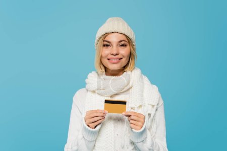 joyful woman in white sweater and winter hat holding credit card isolated on blue