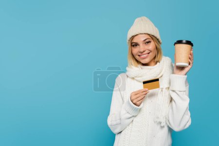 happy blonde woman in white winter outfit holding credit card and paper cup isolated on blue