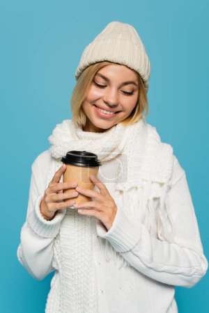 happy blonde woman in white sweater and winter hat holding paper cup isolated on blue