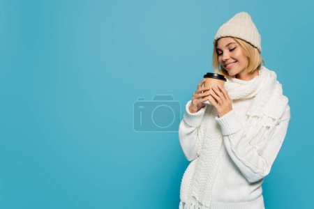 Photo for Happy blonde woman in white sweater and knitted hat holding paper cup with takeaway drink isolated on blue - Royalty Free Image