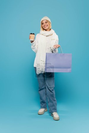 full length of happy blonde woman in white sweater and knitted hat holding paper cup and shopping bag on blue