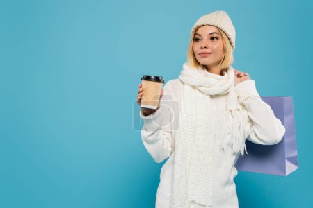 young woman in white sweater and knitted hat holding paper cup and shopping bag isolated on blue