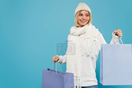 cheerful young woman in white sweater and knitted hat holding shopping bags isolated on blue