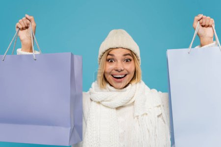 excited young woman in white sweater and knitted hat holding shopping bags isolated on blue