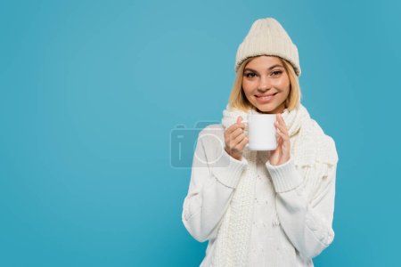 cheerful woman in white sweater and hat holding cup of coffee isolated on blue 