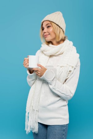 smiling woman in white sweater and winter hat holding cup of coffee isolated on blue 