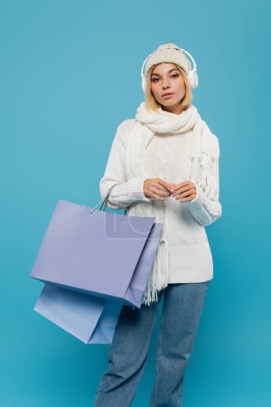 young woman in winter outfit and wireless headphones holding shopping bags isolated on blue 