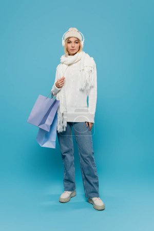 full length of young woman in winter outfit and wireless headphones holding shopping bags on blue 