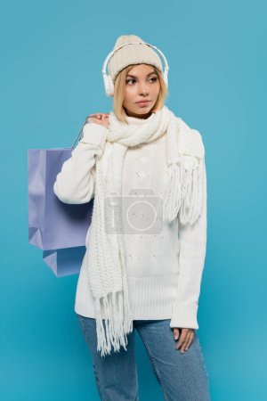 blonde woman in winter outfit and wireless headphones holding shopping bags isolated on blue 