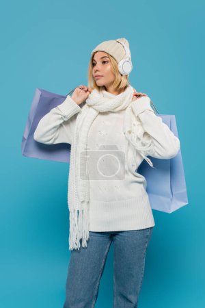 young blonde woman in white winter outfit and wireless headphones holding shopping bags on blue 