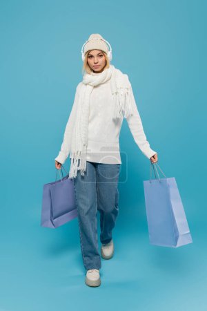 Téléchargez les photos : Pleine longueur de femme blonde en tenue d'hiver et écouteurs sans fil marchant avec des sacs à provisions sur bleu - en image libre de droit