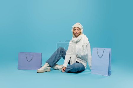 full length of blonde woman in winter outfit and wireless headphones sitting near shopping bags on blue 