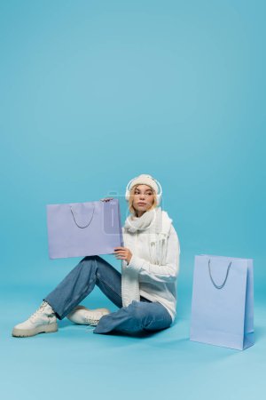 full length of blonde woman in winter outfit and wireless headphones sitting and holding shopping bag on blue 
