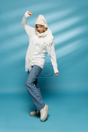 full length of young blonde woman in winter hat and wireless headphones posing on blue 