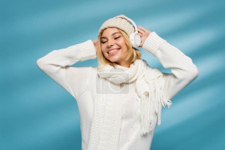 Téléchargez les photos : Femme joyeuse en chapeau d'hiver et écouteurs sans fil écouter de la musique sur bleu - en image libre de droit