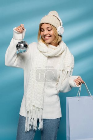 pleased young woman in knitted hat and wireless headphones holding disco ball with shopping bag on blue 