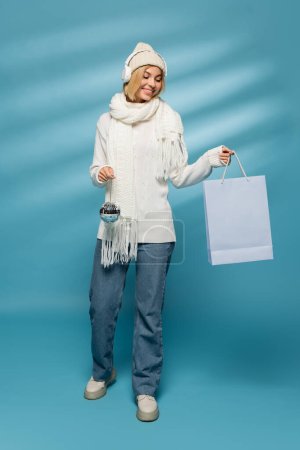 full length of happy young woman in knitted hat and wireless headphones holding disco ball with shopping bag on blue 