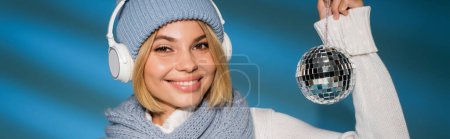 happy woman in winter hat and wireless headphones holding disco ball on blue, banner puzzle 619038952