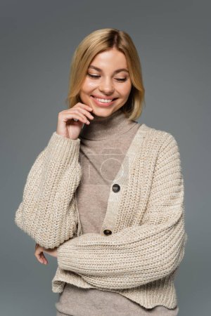 cheerful blonde woman in turtleneck and cardigan smiling isolated on grey