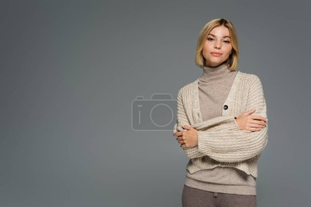 young blonde woman in turtleneck and cardigan standing with crossed arms isolated on grey