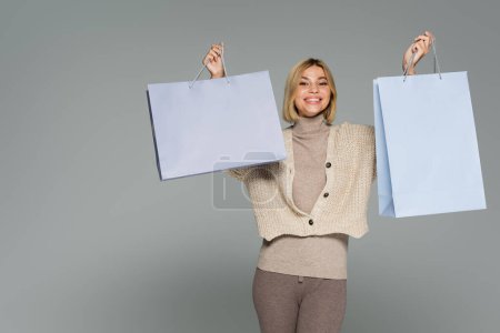 happy blonde woman in turtleneck and cardigan holding shopping bags isolated on grey