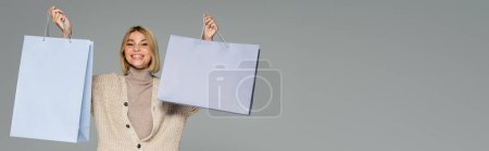 glückliche blonde Frau in Rollkragen und Strickjacke mit Einkaufstaschen auf grau, Banner
