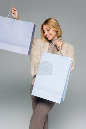 pleased blonde woman in turtleneck and cardigan holding shopping bags on grey