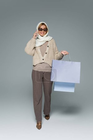 full length of cheerful woman in knitwear and sunglasses holding shopping bags while talking on smartphone on grey