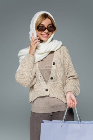 femme joyeuse en tricot et lunettes de soleil tenant des sacs à provisions tout en parlant sur smartphone isolé sur gris