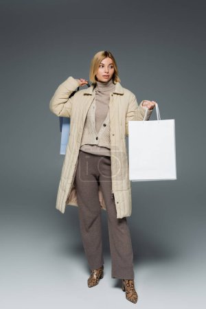 Trendy woman in winter jacket and turtleneck holding shopping bags on grey background 