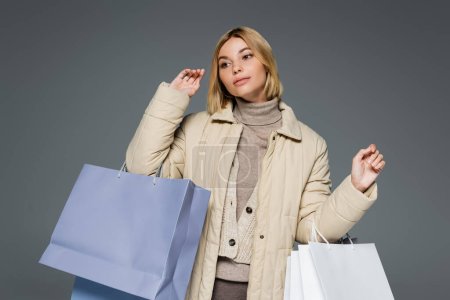 Téléchargez les photos : Femme blonde en tenue d'hiver beige tenant des sacs à provisions isolés sur gris - en image libre de droit