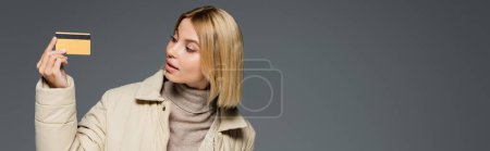 Blonde woman in winter jacket looking at credit card isolated on grey, banner 