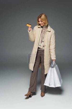 Smiling woman in winter outfit holding credit card and shopping bags on grey background 