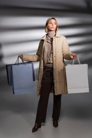 Pleine longueur de jolie femme en tenue d'hiver tenant des sacs à provisions sur fond gris abstrait 