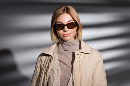Blonde Frau mit Sonnenbrille und Winterjacke auf abstrakt grauem Hintergrund 