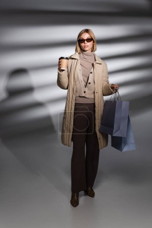 Foto de Mujer joven de moda en chaqueta de invierno celebración de compras y café para ir en fondo gris abstracto - Imagen libre de derechos