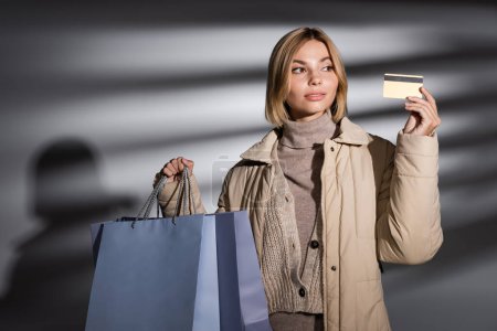 Blonde Frau in Winterjacke mit Einkaufstaschen und Kreditkarte auf abstrakt grauem Hintergrund 