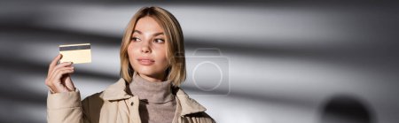 Mujer rubia en chaqueta de invierno con tarjeta de crédito sobre fondo gris abstracto, pancarta 