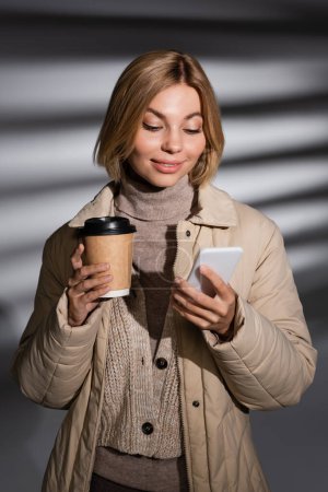 Smiling woman in beige winter jacket holding coffee to go and using smartphone on abstract grey background 