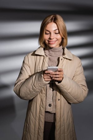 Photo for Positive young woman in winter jacket using cellphone on abstract grey background - Royalty Free Image