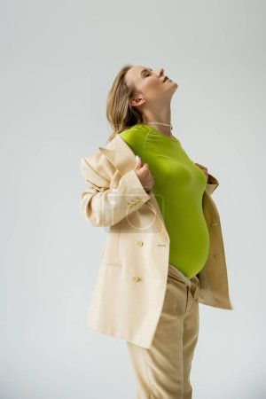 Photo for Stylish pregnant woman touching beige jacket while standing isolated on grey - Royalty Free Image